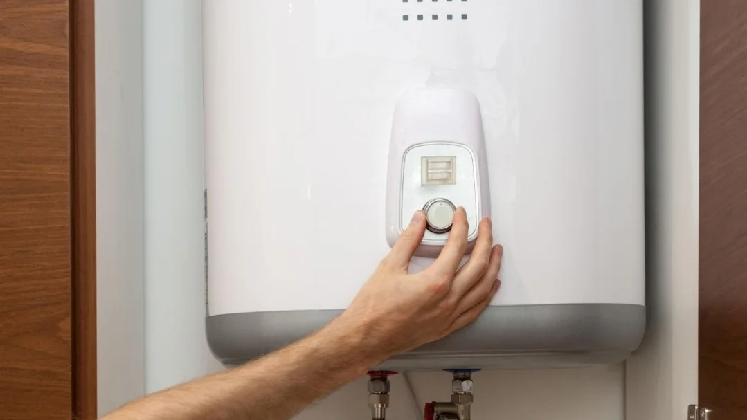 A hand adjusting the thermostat dial on a wall-mounted electric geyser